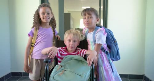 Three school-age children arriving at school doorway, posing around wheelchair and fixing backpacks
