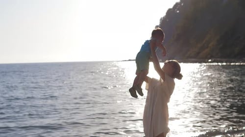 Young Mother of a 2 Years Old Boy is Keeping on Her Arms and Playing with a Son on a Seaside Beach
