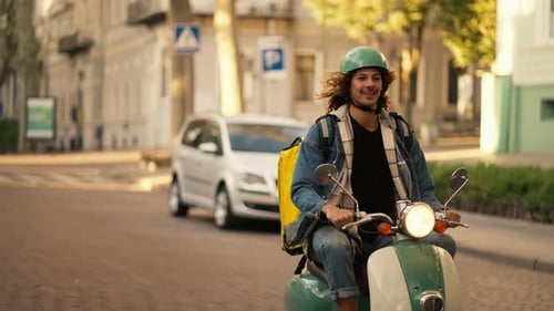 Un gars heureux aux longs cheveux bouclés dans un casque vert et une veste en jean sur un cyclomoteur vert avec un