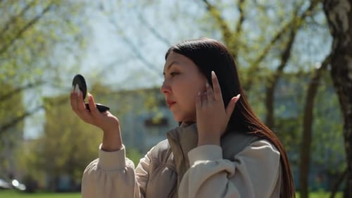 Asian Woman Applying Makeup with Compact Mirror in Park Focused Touchup and Subtle Powder