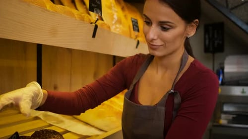 Female baker arranging freshly baked bread on shelves inside bakery.