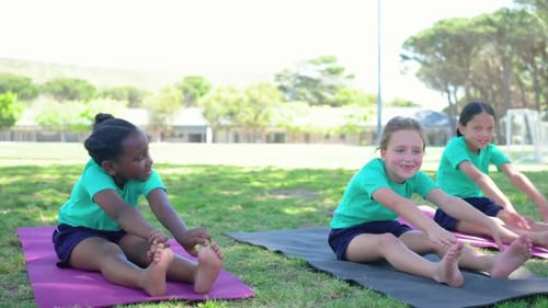 Diverse child girls after coach cue in teal shirts stretching, rotating torsos on mats in park