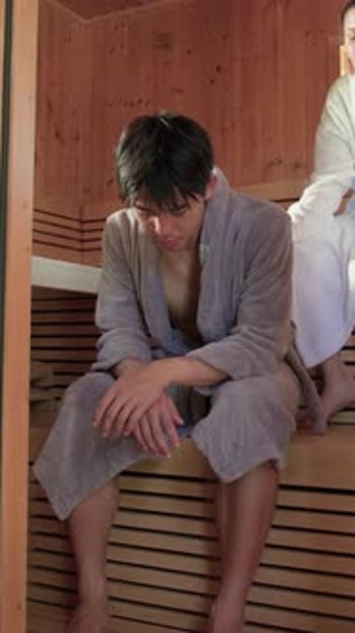 Vidéo verticale : Réagissant à la chaleur du sauna, homme en peignoir gris assis sur un banc en bois se frottant les mains