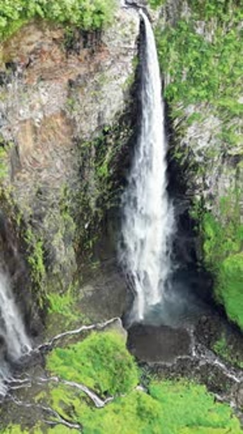 Aerial drone view of multiple narrow waterfalls flowing down steep green cliffs, surrounded by dense