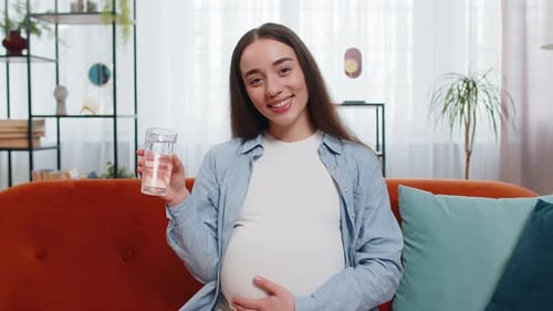 Schwangere Frau, die ein Glas mit Wasser in der Hand hält und trinkt, während sie auf dem Sofa sitzt, gesundes Mutterschaftskonzept
