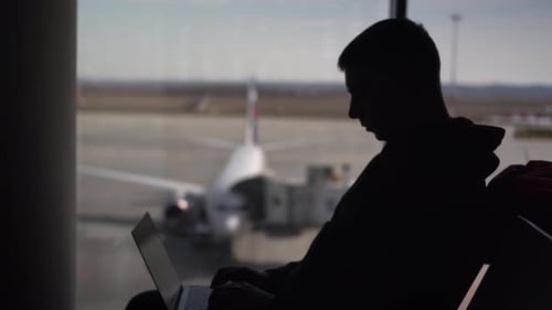 A Young Man in a Sweatshirt is Sitting at the Airport with a Laptop An IT Specialist Works Sitting