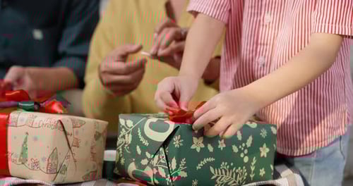 Close-up of young Asian boy wrapping Christmas gift with green and red paper, bow. Child's hands car
