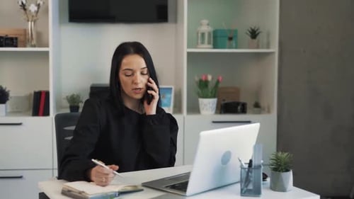 A busy businesswoman sits at a desk in the office and talks on the phone, solves work matters and wr