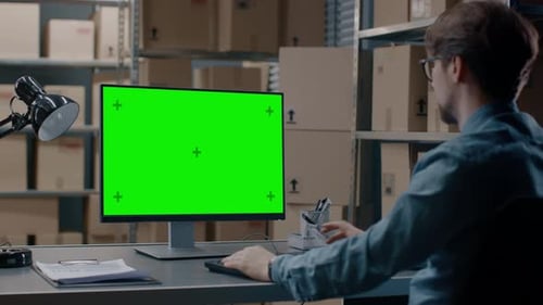 El gerente de inventario del almacén trabaja en una computadora con pantalla de maqueta verde mientras está sentado en su escritorio.
En