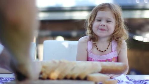 Pre-school girl nodding and smiling as Dad slices bread-stick in backyard picnic.
