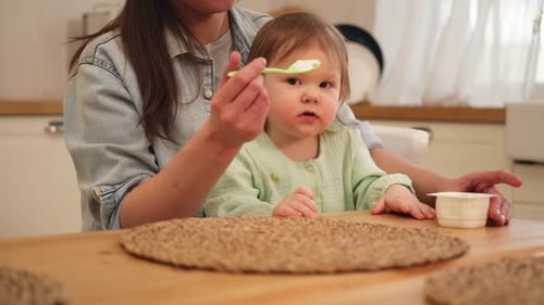 Woman Feeds Child Food in Kitchen at Table