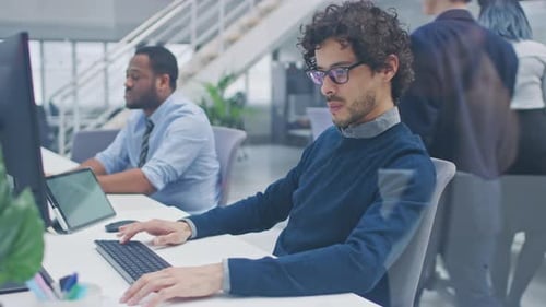 Der junge gutaussehende Manager mit lockigem Haar arbeitet auf einem Desktop-Computer und vergleicht Daten mit einer Software