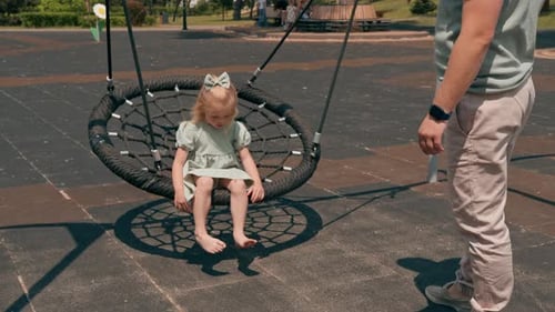 Little cute girl in a green summer dress with a bow on her head on the playground mother rides her