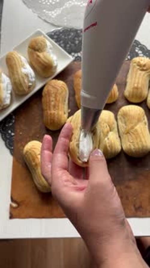 Eine Frau füllt Eclairs mit Sahne und legt sie auf einen Teller. Von hinten geschossen