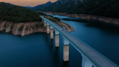 Aerial View of a Car Driving on a Bridge Over a Lake Surrounded By Mountains