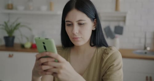 Serious Young Woman Using Smartphone Messaging Looking at Display in Kitchen Alone