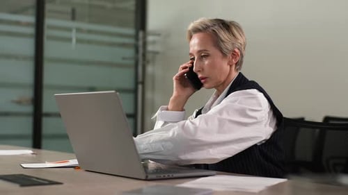 Busy Middleaged Business Woman in Suit Working at Office Sitting on Work Desk with Laptop Holding