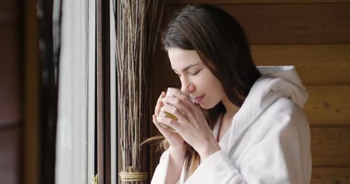 Uma linda mulher olha pela janela usando um roupão branco e bebe chá quente ou chá de ervas l