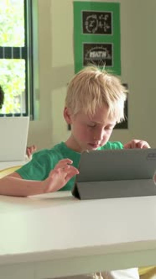 Vertical video: Tapping schoolboy in green tee swiping at school table, studying with tablet folio