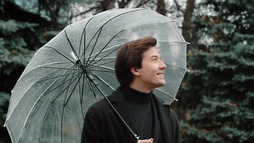 Young Smiling Man in a Coat Standing Under a Transparent Umbrella in the Autumn City Park Looking