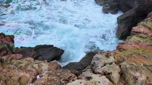 Rocky coastal scene where turbulent white waves crash against jagged, reddish-brown rocks. Foam from