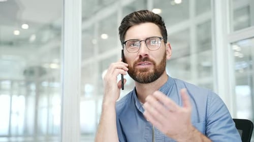 Confident businessman talking on mobile phone while sitting at workplace in modern office. Manager