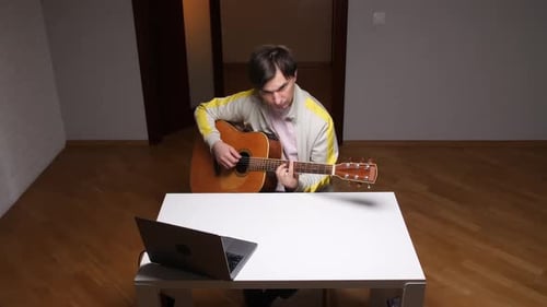 Young Adult Plays Guitar at Table With Laptop