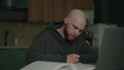 Focused Pensive Male Student Researching Reading Book Taking Notes on Notebook Sitting at Kitchen