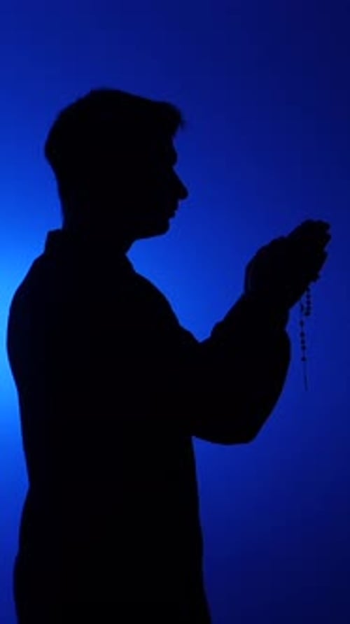 Silhouette of man praying with rosary in blue