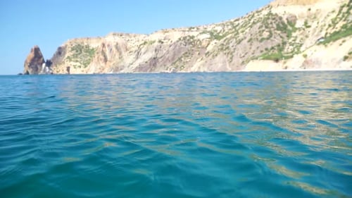 Sea Water Surface Camera Flies Over the Calm Azure Sea with Volcanic Rocky Shores on Background