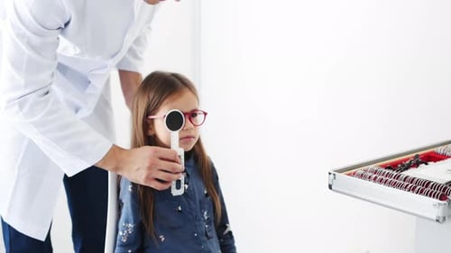 Oculist in white coat wearing glasses on little girl, covering her eyes and cheking her sight