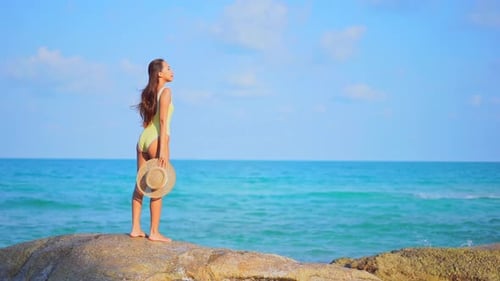 Standing on a large rock a pretty woman in a bathing suit and sun hat in hand looks over the ocean w