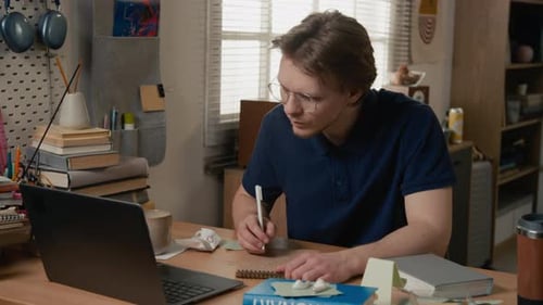 Junger Mann studiert und schreibt in ein Notizbuch, während er online auf dem Laptop sucht