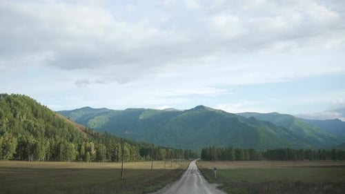 Route rurale pittoresque menant à des montagnes lointaines entourées de champs verts et de forêts sous un ciel nuageux