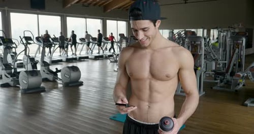 Portrait of a guy in a gym while resting between a workout and the other uses his phone to watch s