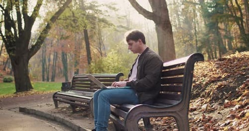 Caucasian Young Man Sitting on Bench in Park and Working on Laptop Outside Male Freelancer Typing on