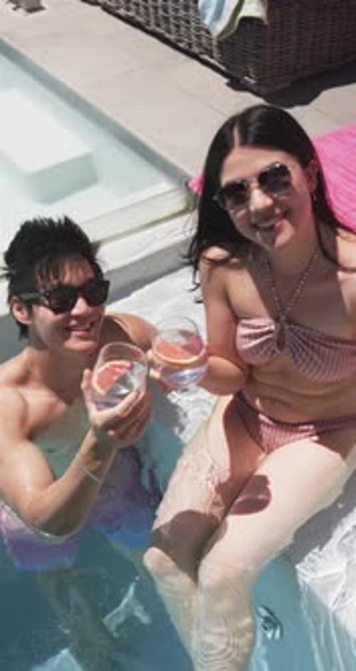 Vertical video: Asian couple holding citrus drinks raising glasses toasting in swimwear at poolside