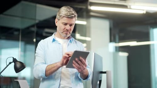 Mature business man uses a digital tablet standing in a modern office indoors. Gray-haired senior