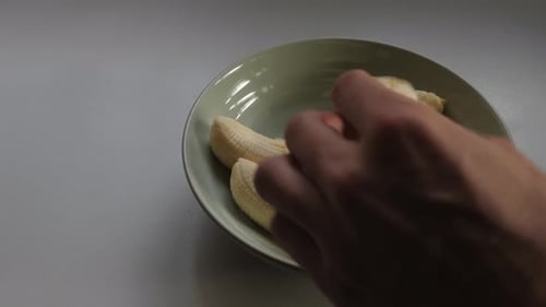 Male hand breaking banana into pieces and putting in small bowl on kitchen bench to make bread