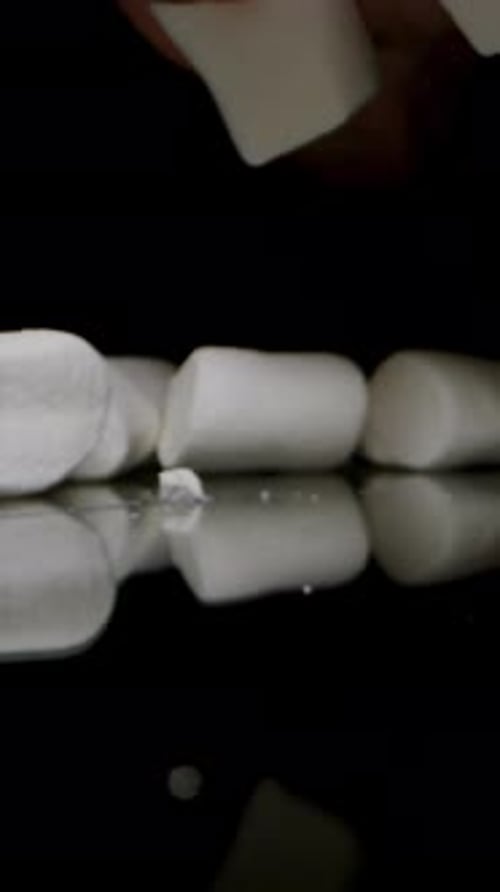 Vertical video. White Marshmallows Thrown onto a Black Table, Hand Appears from Darkness.