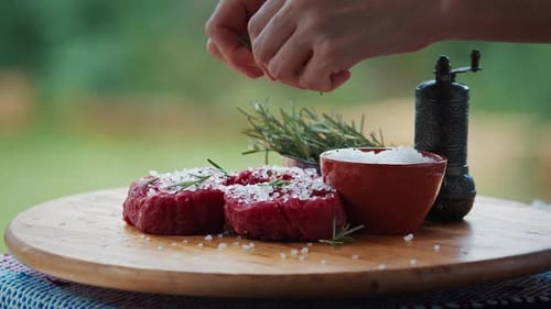 BBQ in the garden. Woman seasons filet mignon resting on the table with pepper and rosemary.