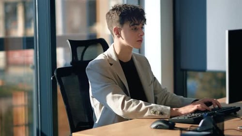 Young man is working in a computer in an office, windows on the background