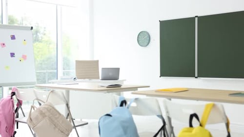 Green chalkboard, backpacks and stylish furniture in classroom. Camera moving right