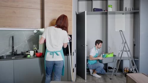 Side View of a Confident Brunette Man in a White Tshirt and Blue Apron Cleaning a Modern Studio