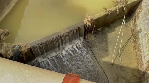 Muddy river water flowing swiftly down a concrete channel, containing floodwaters from heavy winter