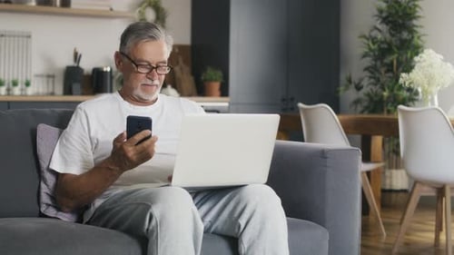 Senior caucasian man sitting at couch and using laptop with mobile phone. Shot with RED helium camer