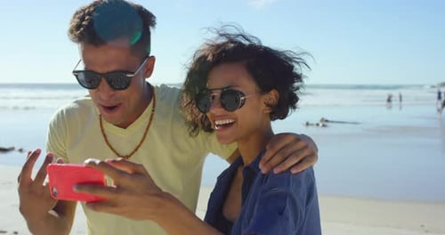 4k video footage of a young couple standing together on the beach and taking selfies on a cellphone