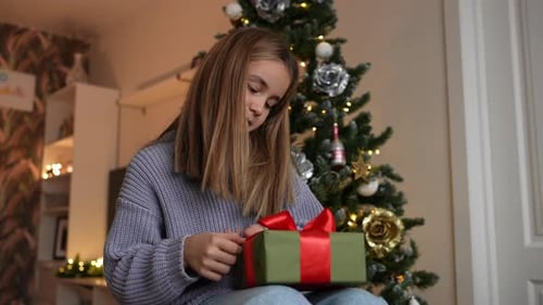 Girl Unwraps Christmas Gift by Christmas Tree