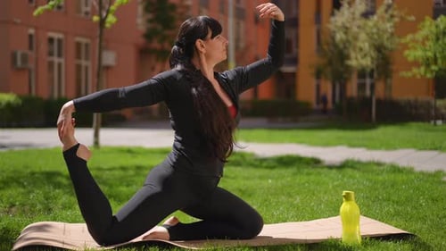 A Woman in a Tracksuit Performs a Yoga Asana While Kneeling on a Yoga Mat Outdoors on a Spring Day