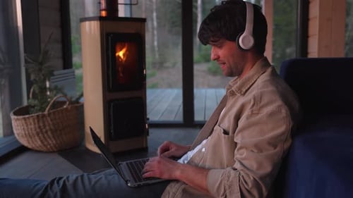 Smiling Man Using a Laptop and Headphones Via Video Call with Friends is Sitting on the Floor in the